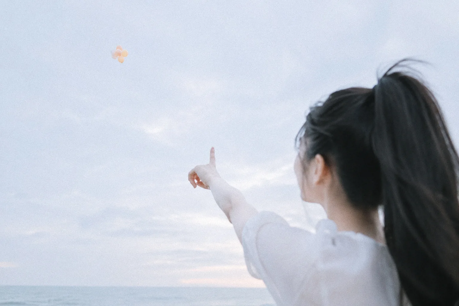 缇薇雅背对镜头望向天空，抬手指向远处的浅色气球